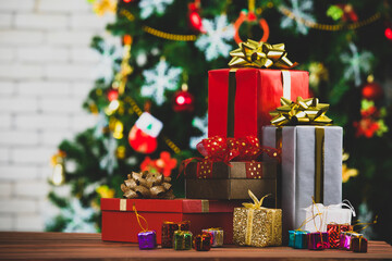 Colorful small and big present gift boxes with shiny ribbon bow tie placed on corner of wooden table in front fully decorative beautiful Christmas eve pine tree and brick wall in blurred background