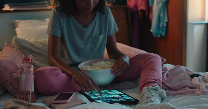 african american woman using tablet computer browsing online social media at home scrolling through picture gallery looking at creative ideas on screen sitting on bed eating popcorn