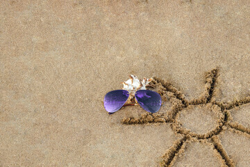 Fototapeta na wymiar Purple sunglasses near shell next to sun pattern on sand. Concept summer, beach holiday. Simple sun drawing in sand on beach, room for your copy space.