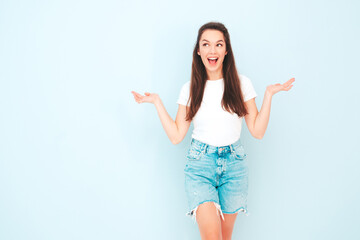 Young beautiful smiling female in trendy summer hipster clothes. Sexy carefree woman posing near light blue wall in studio. Positive model having fun indoors. Raising hands. thinking about choice