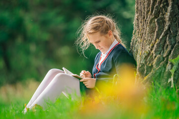 Pretty cute young little school girl in uniform sitting under a tree in park reading book or doing...