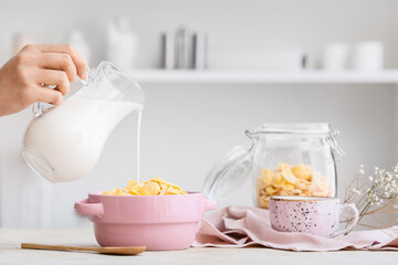 Woman pouring milk from jug into pot with corn flakes on table in kitchen