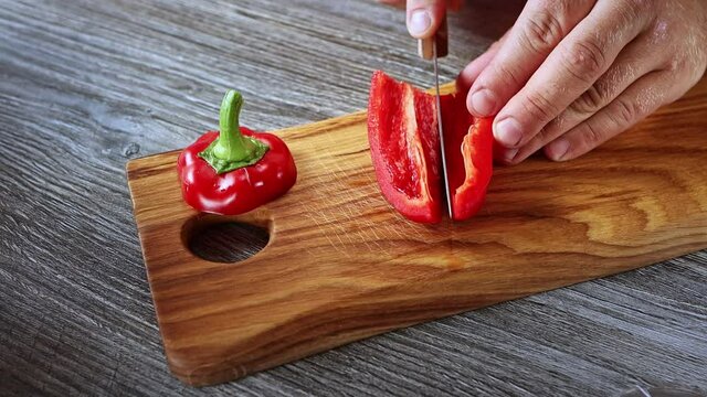 Male hands slowly cutting red bell peper for fresh salad on natural wooden chopping board close up view. Vegetarian food. Helthy food concept. Homemade food cooking. 
