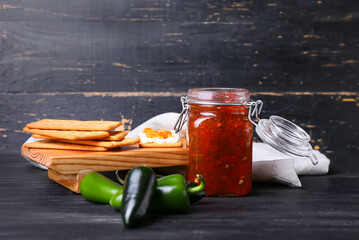 Jar with delicious jalapeno pepper jam and crackers on dark wooden background