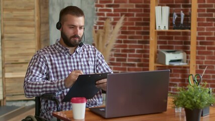 Disabled student sits front of laptop in headset and remotely studies making notes in clipboard, front view. Concept of distance learning