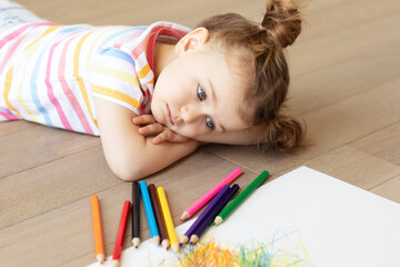 Sad tired frustrated bored stressful child lies on wooden floor with white sheet of paper and colored pencils. Learning difficulties, education concept. Family relationship. Misses home