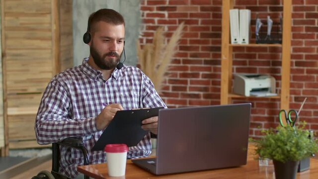 Disabled student sits front of laptop in headset and remotely studies making notes in clipboard, front view. Concept of distance learning