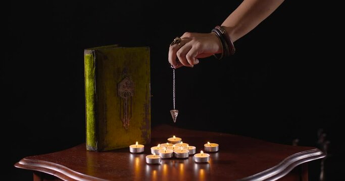 Close-up detailed shot of fortune-teller's hand with pendulum hovering over table with candles and old book on black background. Witch putting spell. Predicting future or Halloween concept