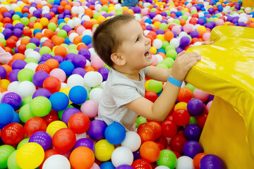 Fototapeta na wymiar Boy kid playing and having a good time in a ball room. Little smiling child playing lying in colorful balls park playground. Happy boy having fun jumping into the ball pit with colorful balls.