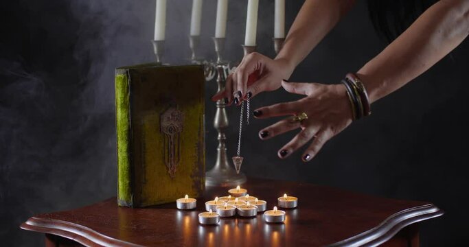 Close-up detailed shot of fortune-teller's hands with pendulum hovering over table with candles and old book on black background with smoke. Witch putting spell. Predicting future or Halloween concept