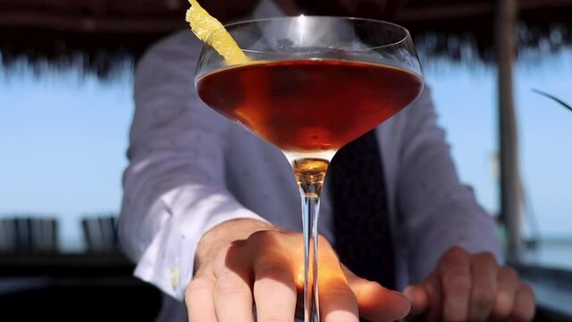 Bartender pushes elegant classic cocktail in coupe glass towards lens in slow-motion at a beachside cabana bar.
