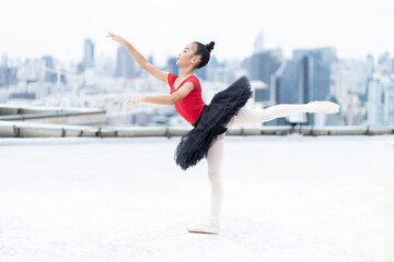 Asian ballerina ballet girl in red, black dress dancing on rooftop with cityscape view background. Young ballerina ballet girl dancing by stretching arms and leg on rooftop