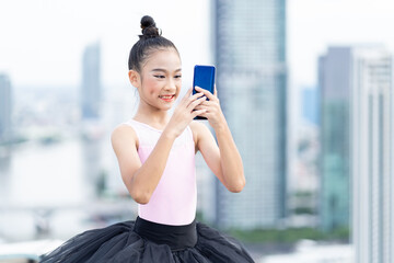 Fototapeta premium Asian ballerina ballet girl take a photo selfie by mobile phone and practicing dancing on rooftop and skyscraper city view background. Female ballet dancer practicing dancing.