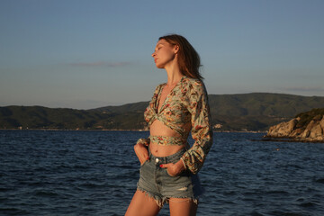 Outdoor portrait of young fashionable woman in crop top and denim shorts with blue sea and clear sky behind. 