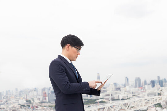 Asian Businessman In Formal Suit Using Digital Tablet On Rooftop With Cityscape View Background