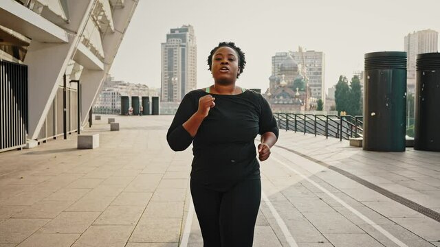 Hard slimming process. Outdoor portrait of young overweight african american woman running alone in morning