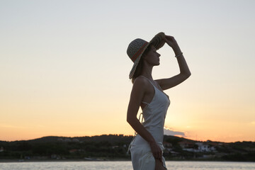 Fototapeta premium Young woman in long white dress and straw hat on the beach during sunset. Stylish summer vacation fashion.