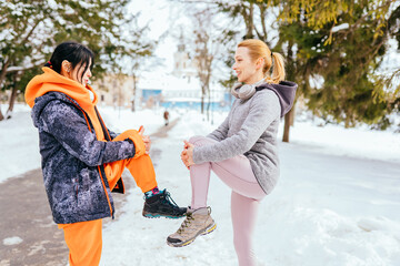 Two shape fitness women in warm sportswear doing leg stretching with her personal trainer in the city park in the sunny winter morning.