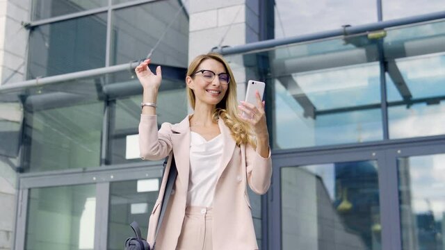 Extremely happy positive business woman in stylish suit dancing on street near office building, holding smartphone in hand, celebrating project approval, receiving permission on company work, success