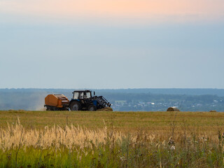 Tractor collects hay in round bales on a summer agricultural field, against the background of the hills and the cloudy sky. Harvesting in the fields. Nature of Russia.