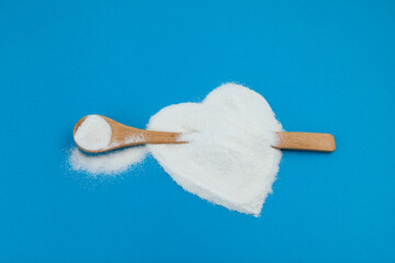 White powder is scattered on blue surface in shape of heart. Wooden spoon with Collagen or protein. Healthy food supplement