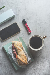 Sandwich lunch or breakfast on gray desk with coffee, smartphone and cosmetic , top view.