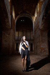 Fototapeta na wymiar A brunette girl with long hair in an evening dress in a vintage Gothic castle. A woman who looks like an evil witch or sorceress in an abandoned old church. Female Model posing in Halloween