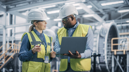 Two Diverse Professional Heavy Industry Engineers Wearing Safety Uniform and Hard Hats Working on Laptop Computer. African American Technician and Female Worker Talking on a Meeting in a Factory. - obrazy, fototapety, plakaty