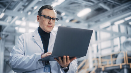 Professional Male Heavy Industry Engineer/Worker Working on a Laptop Computer. Confident Caucasian Industrial Specialist Wearing White Laboratory Robe in a Factory Facility.