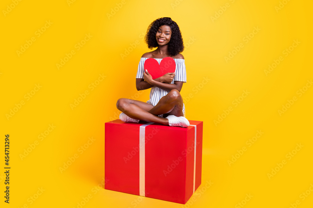 Wall mural Portrait of beautiful trendy cheerful girl sitting on big giftbox hugging paper heart card isolated over bright yellow color background