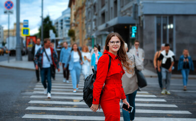 Lovely joyful girl in glasses and red overalls standing with mobile phone at crowded crosswalk