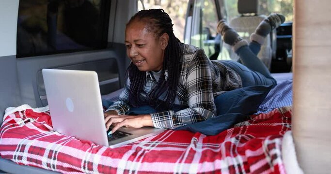 Mature african woman using computer laptop inside mini van camper - Elderly person on a road trip vacation