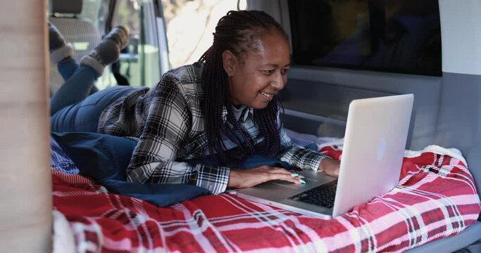 Mature african woman using computer laptop inside mini van camper - Elderly person on a road trip vacation
