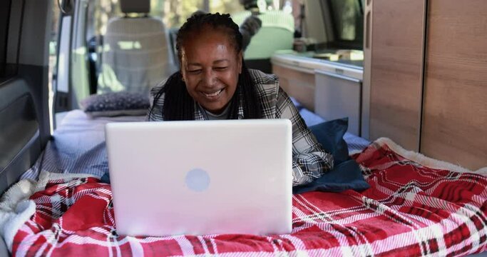 Mature african woman using computer laptop inside mini van camper - Elderly person on a road trip vacation