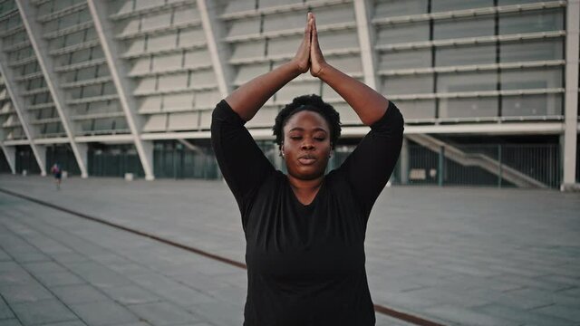 Zoom out portrait of mindful chubby african american woman meditating outdoors, practicing breathing exercise