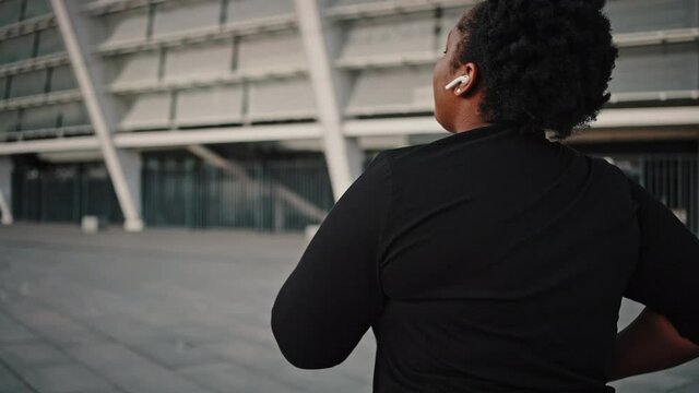 Overweight african american woman practicing run workout outdoors, enjoying music in wireless earphones, tracking shot