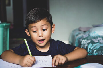 The Asian boy sitting behind the desk was writing something with an excited expression. Study at...