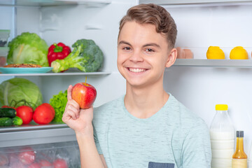 Smiling handsome young teen boy holding fresh red apple while standing near open fridge in kitchen at home. Portrait of pretty child choosing food in refrigerator full of healthy products - 454719419