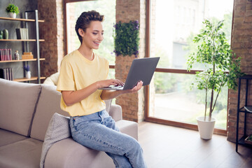 Photo of pretty sweet young lady wear yellow t-shirt sitting sofa typing modern gadget indoors room home