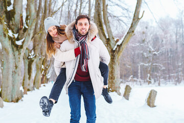 Happy young man carry his smiling pretty woman on his back at winter park. Boyfriend giving...