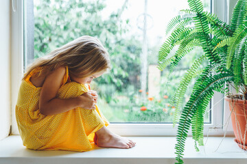 Five years old blonde girl staring out of the window on a wet cold rainy day. Sad child look to...