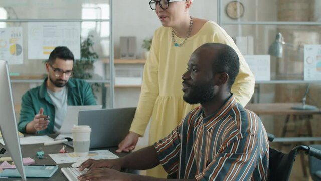 Tilt up shot of Afro-American businessman with disability in wheelchair typing on keyboard and discussing project on computer with Caucasian female colleague while cooperating in office