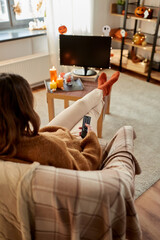 halloween, holidays and leisure concept - young woman with remote control watching tv and resting her feet on table at cozy home