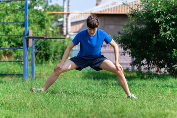teenage boy exercising outdoors, sports ground in the yard, he squats and does a warm-up, healthy...