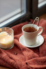 season, leisure and objects concept - cup with mesh tea infuser ball, earrings and candle on window sill at home