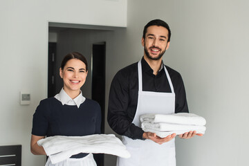 smiling housekeepers holding white bed sheets and towels in hotel room