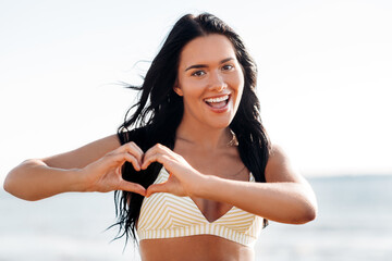 people, summer and swimwear concept - happy smiling young woman in bikini swimsuit showing hand heart gesture on beach
