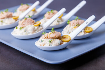 Cold appetizer made from cod liver, cod caviar, olives, cucumber and microgreens on serving spoons. Traditional cold dish. Close-up, blue plate, black background.