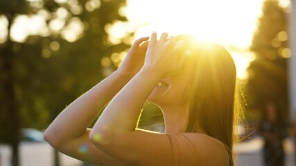 Cheerful woman wearing summer top standing near building and touching hair in rays of sunset, blurred background. Arc shot portrait of young female. Concept of happiness