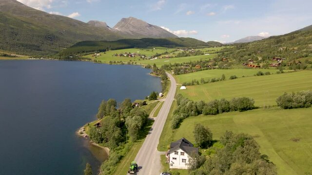 Cars Driving On The Road By The Lake Shore Of Eidsvatnet In Eidsdal, Norway With Mountain Village Of Aarset In Background. aerial sideways
